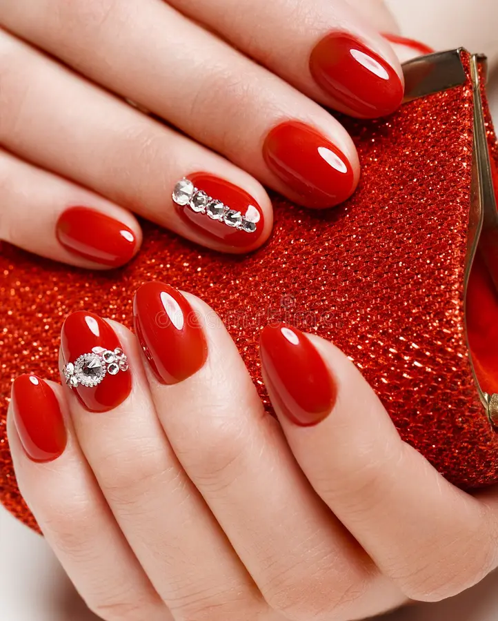 Uñas ovaladas en rojo intenso y brillante con acentos de cristales, sobre un clutch rojo escarchado